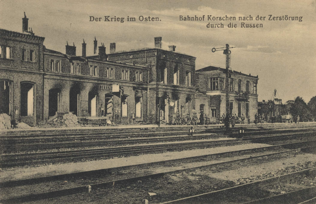Schwarz-weiß-Fotografie des Bahnhofs Korschen in Ostern, Deutschland, mit Bahnschienen, Bahnhofsgebäuden, Menschen, Bäumen und Himmel.
