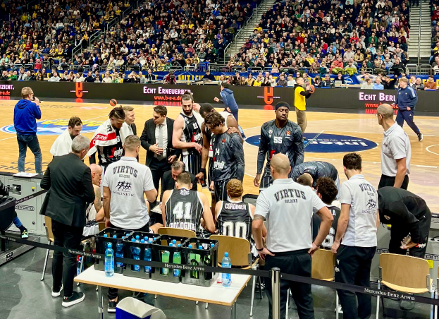 Eine Gruppe von Männern steht auf einem Basketballfeld umgeben von Zuschauern, mit einer Person, die eine Kamera auf der linken Seite hält und Tafeln mit Text im Hintergrund.