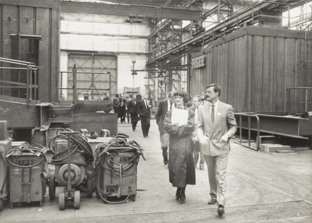 Gruppe von Menschen in Anzügen, die durch eine industrielle Fabrik gehen, mit Maschinen auf der linken Seite und einer Wand im Hintergrund.