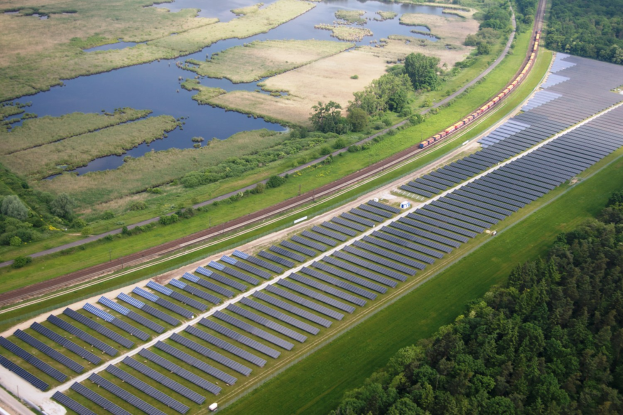 Luftaufnahme einer Solar-Farm mit Panelen, umgeben von Bäumen, Gras und Wasser, mit einem Zug auf einer nahen Bahnstrecke.