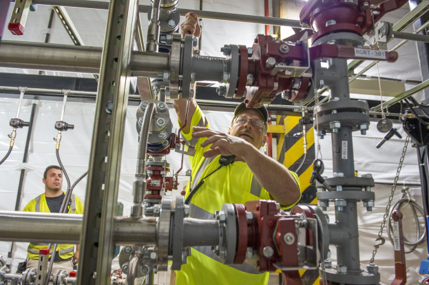 Zwei Männer in gelben Sicherheitswesten und Jacken arbeiten an einer Maschine in einer Fabrik mit Metallstangen, Rohren und einem weißen Tuch im Hintergrund; einer trägt eine Brille und eine Kappe.