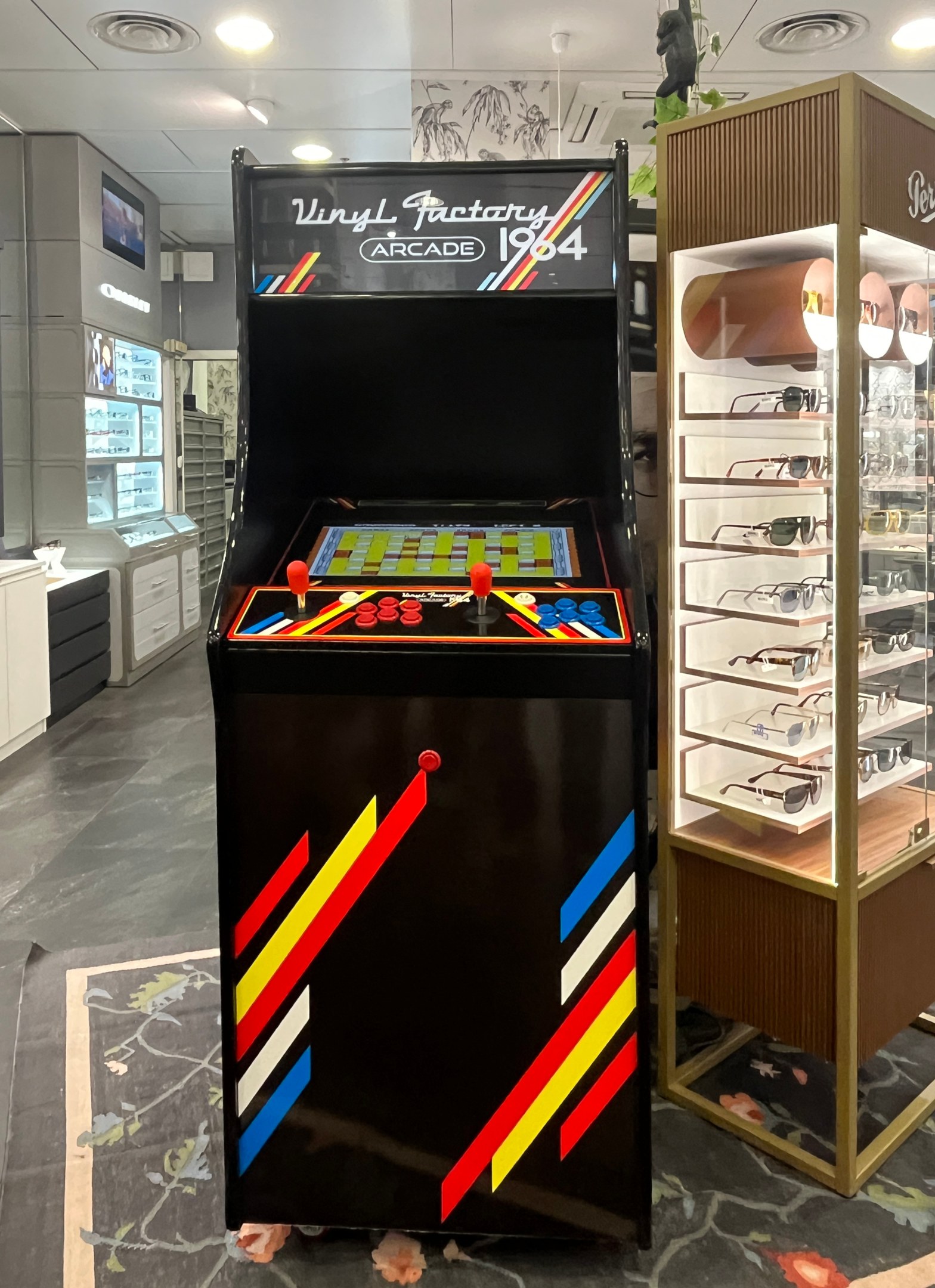 Ein Geschäft mit einem zentralen Retro-Arcade-Automaten, einer Glasvitrine auf der rechten Seite und einer Retro-Beleuchtung mit Schränken, einem Fernseher und anderen Gegenständen im Hintergrund.