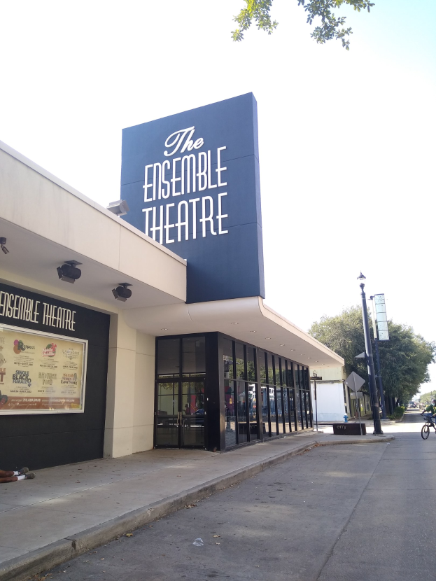 Außenansicht des Ensemble Theaters in San Francisco mit Glaswänden, einer sitzenden Person auf dem Gehweg, einem Fahrrad, einer Hinweistafel, Straßen-Elementen, Bäumen und einer bewölkten Himmel.