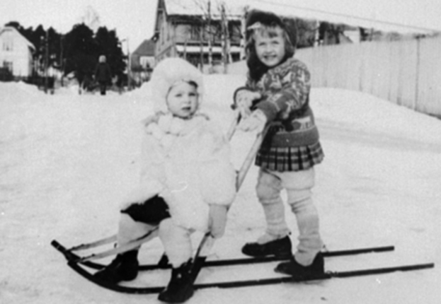 Zwei junge Mädchen auf Skiern im Schnee, eine sitzt auf einem Schlitten, mit Häusern, Bäumen und einer Wand im Hintergrund, schwarz-weiß.