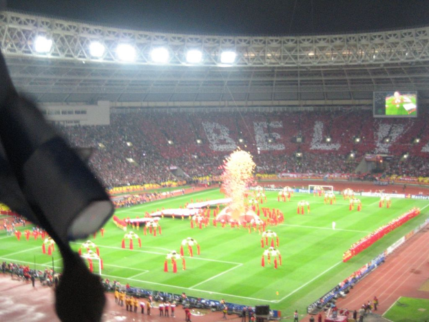Ein Stadion mit Flutlicht, Eisengittern, Zuschauern, stehenden Individuen, Werbetafeln, Anzeigetafeln, Sicherheitskameras, Fahnen und Dekorationen.