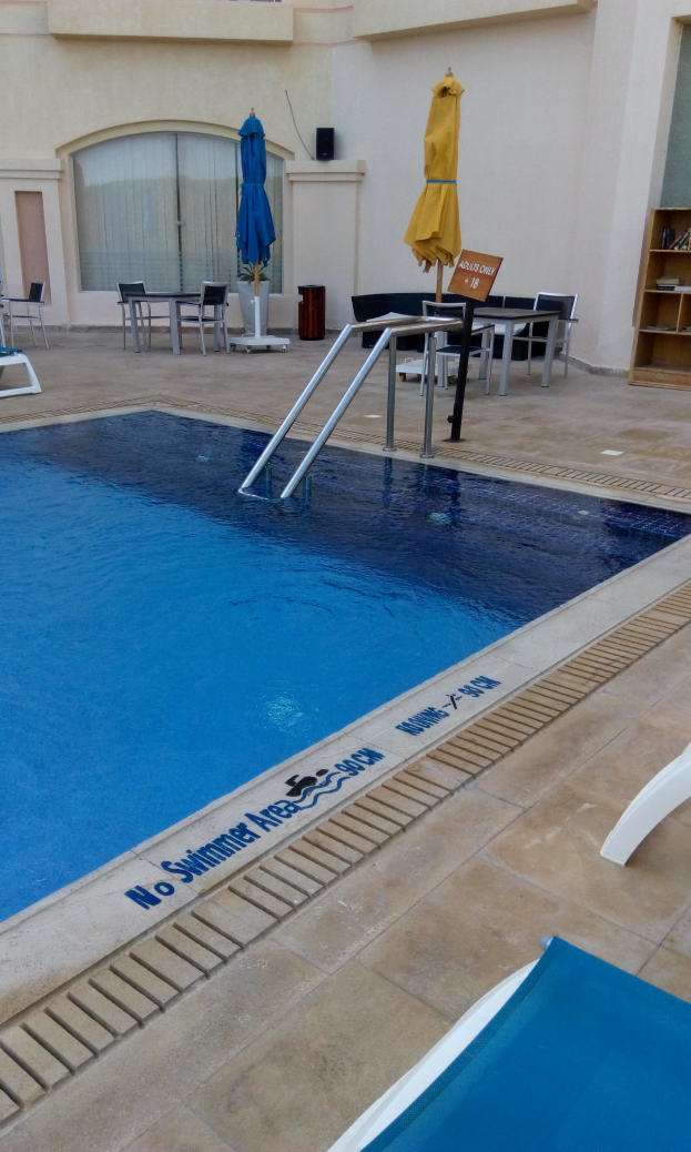 Ein Schwimmbad mit Liegestühlen, Sonnenschirmen, Tischen und Stühlen, mit einem Gebäude im Hintergrund.