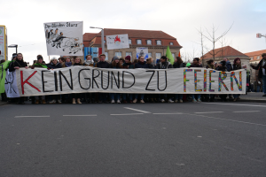 Demonstranten mit einem Transparent mit der Aufschrift 'Kein Grund zur Feier' gegen deutsche Sparmaßnahmen, mit Gebäuden, Laternen, Bäumen und einem klaren Himmel im Hintergrund.
