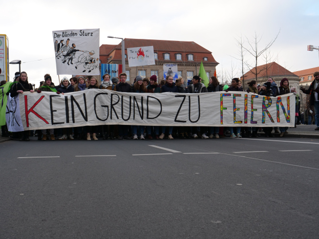 Demonstranten mit einem Transparent mit der Aufschrift 'Kein Grund zur Feier' gegen deutsche Sparmaßnahmen, mit Gebäuden, Laternen, Bäumen und einem klaren Himmel im Hintergrund.