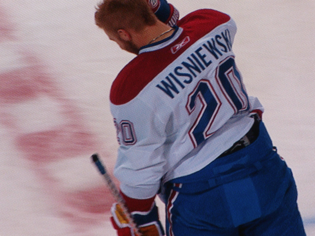 Ein Eishockeyspieler in einem "Wisniewski"-Trikot steht auf dem Eis und hölt einen Eishockeystock, mit einem konzentrierten und entschlossenen Gesichtsausdruck.