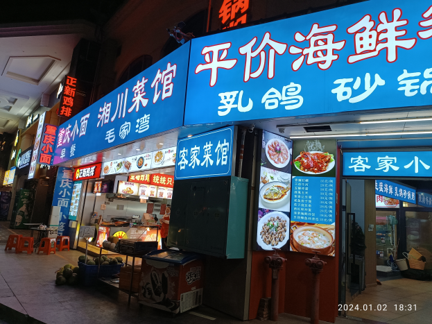 Ein chinesisches Restaurant an einer Straßenecke bei Nacht mit Tischen, Hockern und anderen Gegenständen auf dem Gehweg, neben einem Geschäft mit einer Glastür, die Lebensmittel und andere Waren zeigt, mit einem Wasserzeichen auf dem Bild.