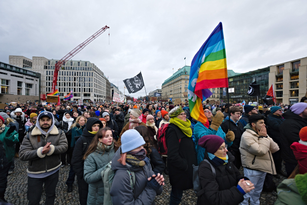 Eine große Gruppe von Menschen mit Fahnen und Schildern vor einem Gebäude während einer LGBTQ+-Rechtsdemo in Berlin, mit Gebäuden, einem Kran und Wolken im Hintergrund.