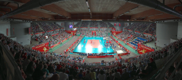Eine große Halle voller Zuschauer bei einem Volleyballspiel mit Bannern, Bildschirmen und Deckenleuchten sowie Geländern unten.