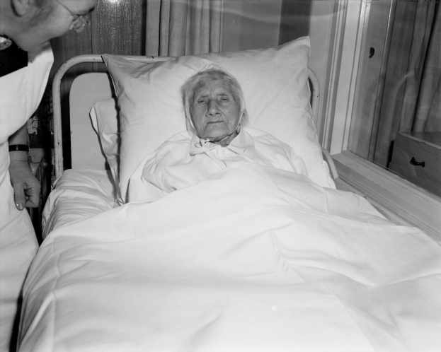 Alte Frau im Krankenhausbett mit einer Krankenschwester daneben, Bett mit weißer Bettdecke und Kissen bedeckt, Fenster mit Vorhängen im Hintergrund.