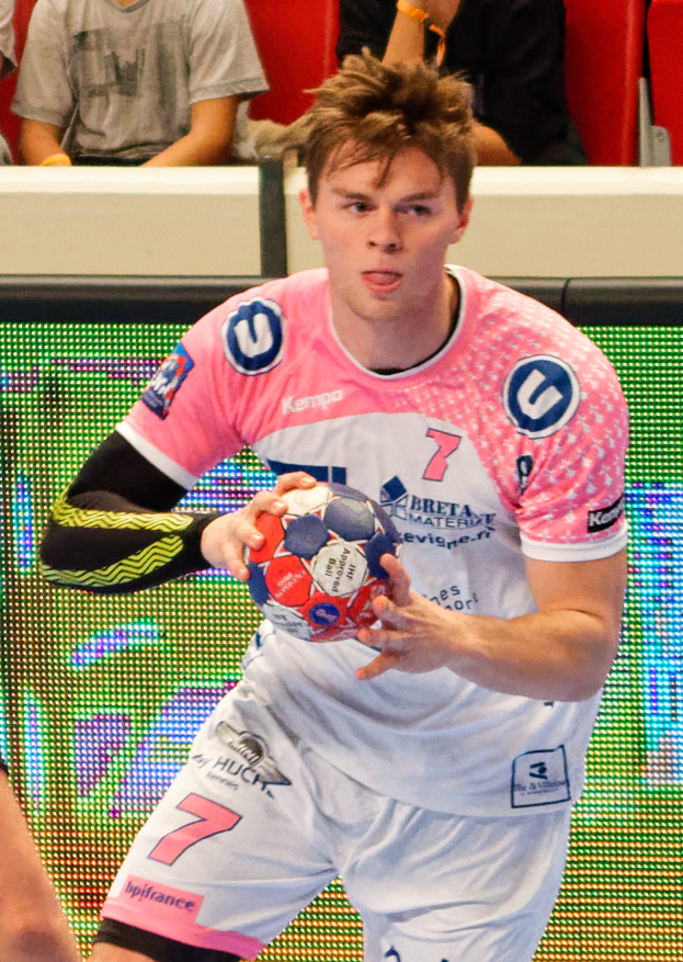 Ein Mann in einem pinken Hemd hält einen Fußball, mit der Hand eines anderen Menschen auf der linken Seite und einigen Leuten im Hintergrund auf Stühlen sitzend.