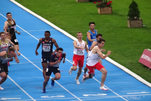 Eine Gruppe von Männern, die auf einer blauen Bahn laufen, umgeben von grünem Gras und Blumentöpfen, mit einer Texttafel auf der rechten Seite, die auf einen Wettbewerb hinweist.