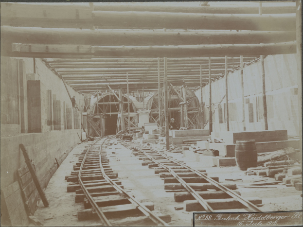 Schwarz-weiß-Foto einer Bahnschiene in einem Gebäude unter Bau, mit verstreuten Holzgegenständen, Fässern, Wänden, Pfählen und Text am unteren Rand.