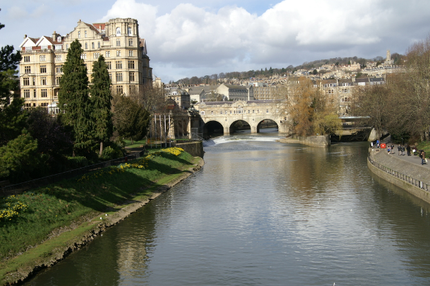 Eine Gruppe von Menschen, die auf einer Straße neben Bäumen und Wasser gehen, mit Gebäuden, Pfählen und einer Brücke im Hintergrund.