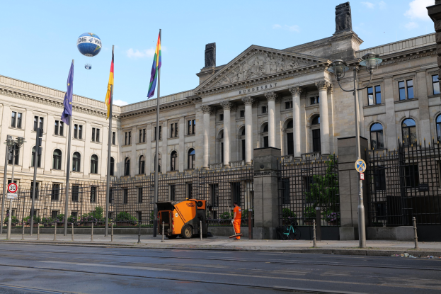 Großes modernes Regierungsgebäude mit vielen Fenstern, umgeben von Straßenmöbeln, Fahrzeugen, Passanten und Bäumen, unter einem bewölkten Himmel.