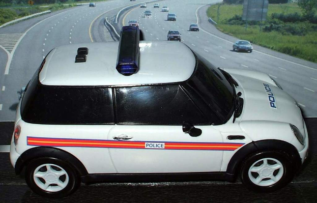 Ein weißes Polizeiauto steht im Vordergrund mit mehreren Autos, die sich auf der Straße dahinter bewegen, umgeben von Gras und Bäumen auf beiden Seiten, in einer ländlichen Umgebung.