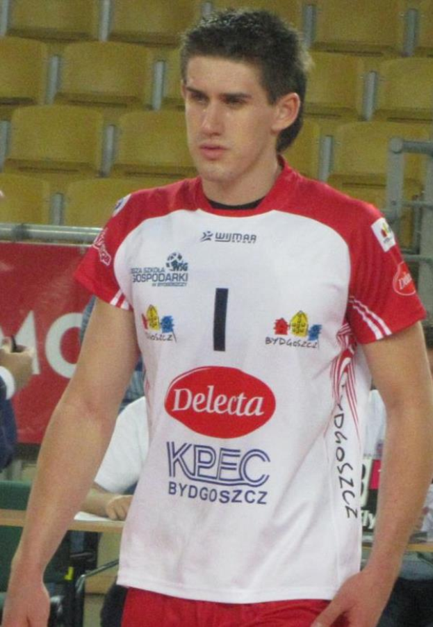 Ein Mann in rot-weißer Handballuniform steht auf einem Feld, umgeben von Zuschauern, mit einem roten Banner im Hintergrund, auf dem 'Krasnoyarsk vs Bydgoszcz' steht.