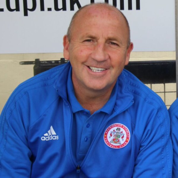 Zwei Männer in blauen Jacken sitzen neben einem anderen Mann, der neben einer Wand mit einer Tafel steht. Der Mann in der blauen Jacke wird als Trainer des Accrington Stanley Football Club identifiziert.