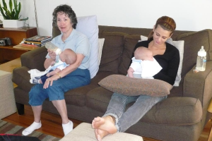 Zwei Frauen sitzen auf einem Sofa und halten Babys, mit Kissen drumherum, vor einer weißer Wand, mit einem Schreibtisch, Bürchern und einer Zimmerpflanze links daneben.