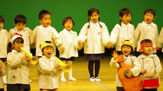 Kinder stehen auf einer B√ľhne und singen mit einem gr√ľnen Vorhang im Hintergrund.
