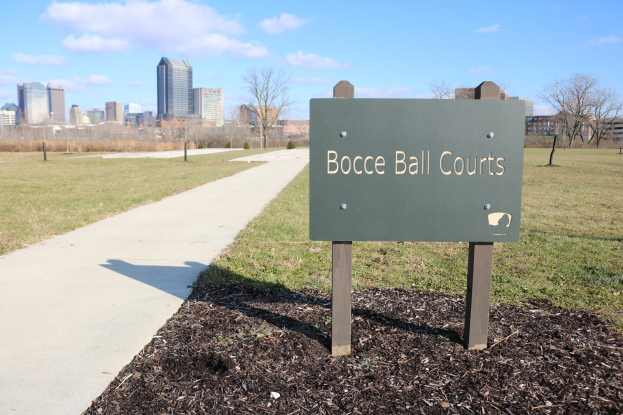 Ein Schild am Boden neben einem Weg und Bäumen trägt die Aufschrift "Bocce Ball Courts in Indianapolis, Indiana" mit einem bewölkten Himmel im Hintergrund.