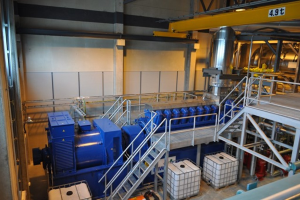 Ein Fabrikinneres mit Trägern, einer Treppe, einem Geländer, Containern, Lampen, Rohren und Wänden.