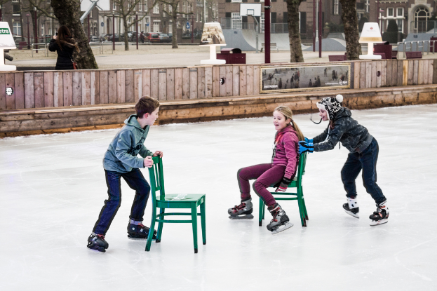 Kinder beim Skifahren vor einem Spielplatz mit drei Kindern und zwei Stühlen in der Mitte und Gebäuden, Bäumen, Bänken, Pfählen und einem Basketballfeld im Hintergrund.