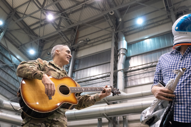 Ein Mann in Militäruniform spielt eine Gitarre, whrend er neben einem anderen Mann in einem Helm steht, mit Rohren und Lichtern im Hintergrund.