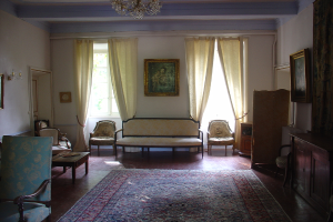 Ein Wohnzimmer mit einer Sitzgruppe, einem Foto auf dem Wand, einem Fenster mit Vorhängen, einer Deckenlampe, Stühlen und einem Tisch mit einem Buch links und einer Kommode, einem weiteren Foto, einer Tür und einem weiteren Fenster mit Vorhängen rechts.