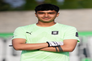 Ein junger Mann in einem grünen T-Shirt steht auf einem Fußballfeld und schaut mit verschränkten Armen und einem Lächeln in die Kamera, während der Hintergrund unscharf ist und darauf hindeutet, dass er im Fokus der Aufnahme ist.