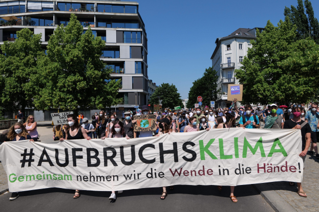 Eine Gruppe von Menschen mit Masken hält ein Banner mit der Aufschrift "Aufbruchsklima" vor einem Gebäude, umgeben von Bäumen und einem klaren Himmel.
