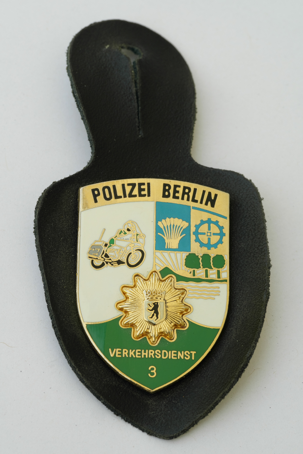 Ein Polizeistern mit der Aufschrift "Polizei Berlin" darauf, der ein blaues Schild mit einem weißen Stern in der Mitte zeigt, umgeben von einem weißen Rand, auf einer Oberfläche platziert.