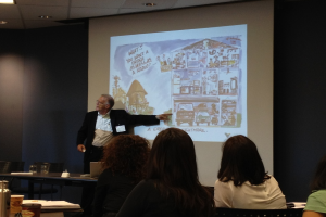 Ein Mann in einem weißen Hemd und schwarzem Blazer zeigt auf ein Fahrzeug auf einem Bildschirm in einem Konferenzsaal, mit drei Frauen im Vordergrund und einem Tisch mit einer Wasserflasche, einem Glas und einem Buch links.