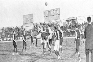 Ein Schwarz-Weiß-Bild einer Gruppe von Männern, die auf einem Feld Fußball spielen, mit einem Ball in der Luft und Zuschauern im Hintergrund, mit Tafeln, auf denen wahrscheinlich text zum Spiel steht.