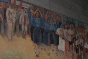 Eine Gruppe von Menschen mit blau gekleideten Mädchen in der Mitte, die klatschen, Musiker auf der rechten Seite, Kinder, die in der rechten Ecke spielen, und Individuals mit roten Fahnen auf der linken Seite.