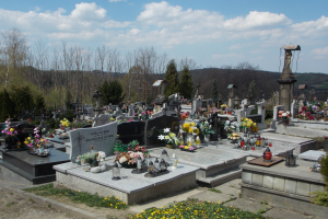 Ein Friedhof mit zahlreichen Grabsteinen, geschmückt mit Blumensträußen, Kerzen und anderen Gegenständen, vor einem Hintergrund aus Bäumen, Hügeln und einem bewölkten Himmel.