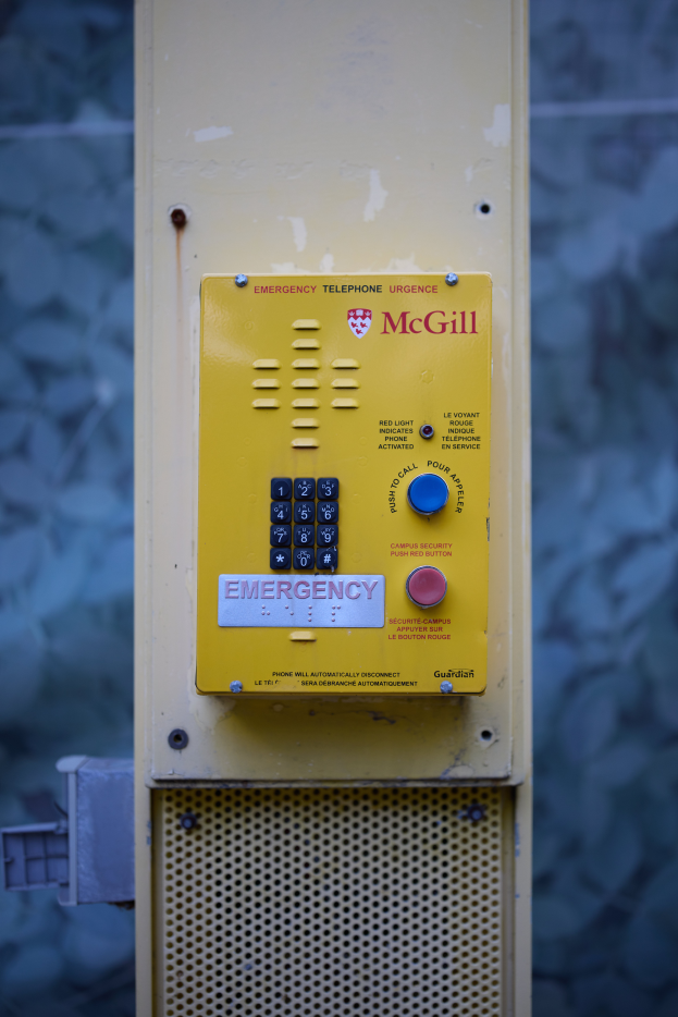 Ein gelbes Notruf-Telefon an der Seite eines Gebäudes angebracht, mit Tasten und Text zum Absetzen von Notrufen, vor einer Wand im Hintergrund.