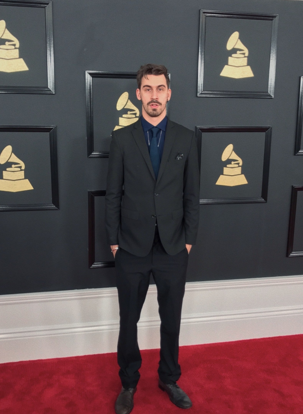 Ein Mann in einem Anzug steht auf dem roten Teppich bei den 55. Annual Grammy Awards, umgeben von einer Wand mit gerahmten Fotos von der Veranstaltung.