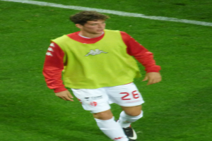 Ein Mann in einem gelb-roten T-Shirt läuft auf einem Fußballfeld, das mit weißen Linien markiert ist und trägt Fußballschuhe.