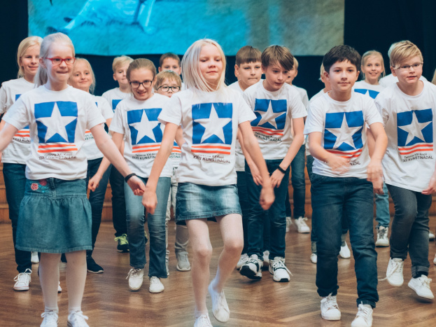 Gruppe von Kindern in weißen Hemden und blauen Shorts, die auf einem Holzfußboden während eines Kunstnädler-Tanzwettbewerbs tanzen, mit einem Bildschirm an der Wand im Hintergrund.