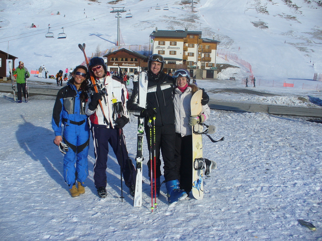 Vier Personen posieren für ein Foto mit Skate-Pads und Werkzeugen, mit Gebäuden und anderen Menschen im Hintergrund, vor einer schneebedeckten und vereisten Berglandschaft.