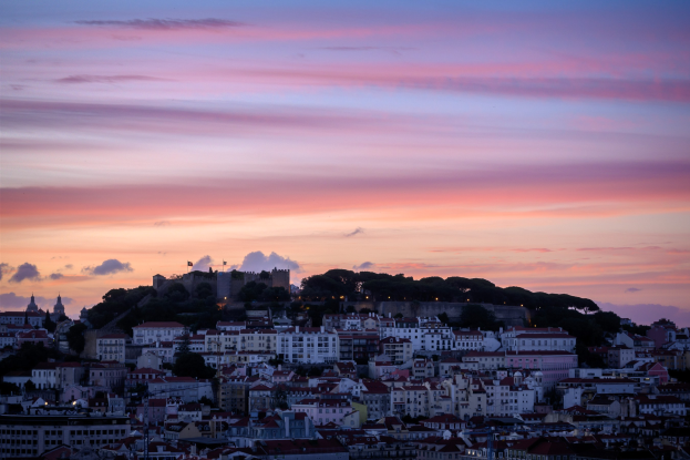 Sonnenuntergangsblick auf Lissabon, Portugal, von einem Hügel aus, mit Gebäuden und Bäumen im Vordergrund und Wolken am Himmel.