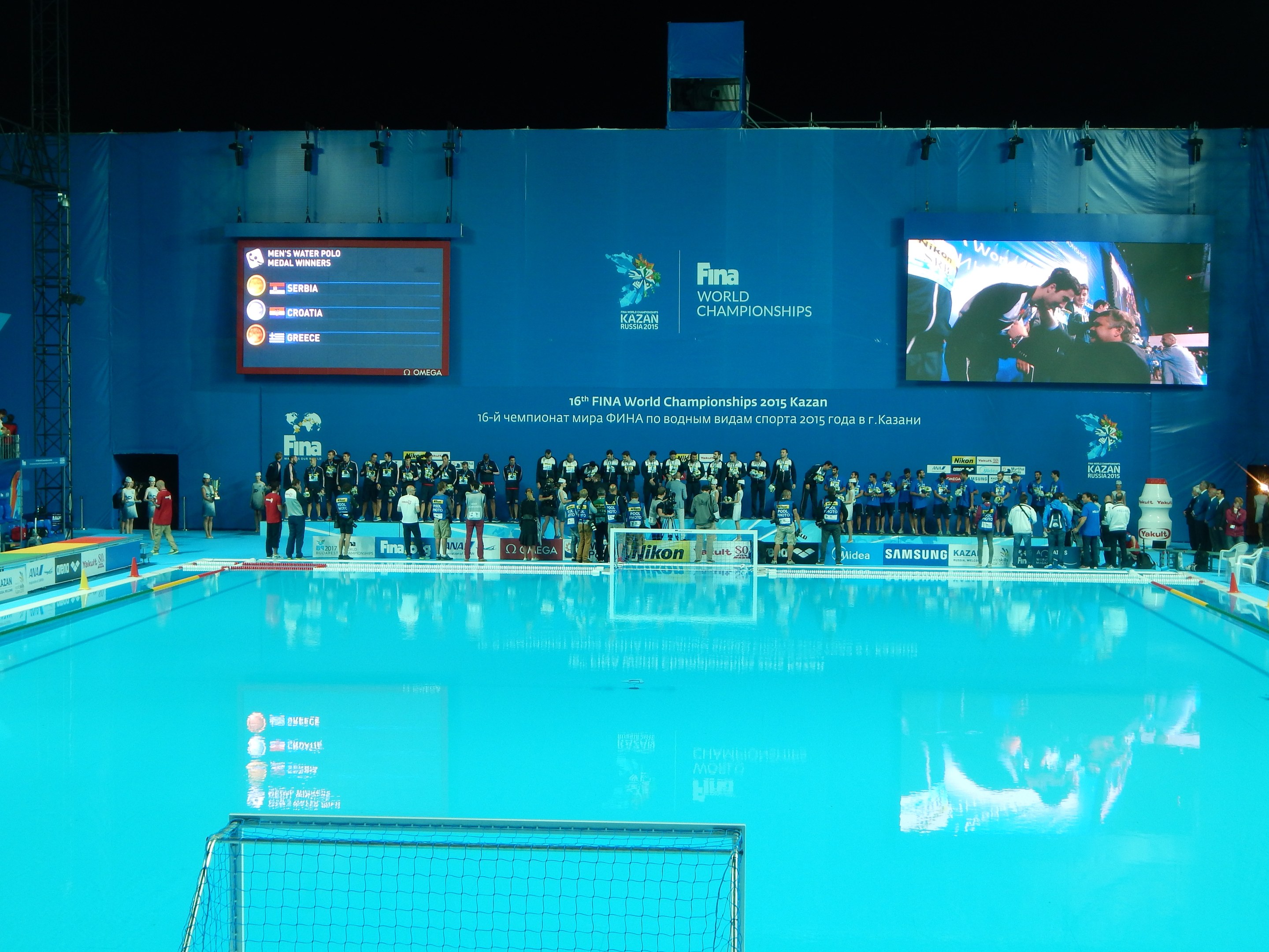 Eine Gruppe von Menschen steht um einen Swimmingpool mit einem Netz am Boden, mit einer Wand im Hintergrund, die "FINA World Championships 2015 Kazan"-Text und Logos anzeigt, sowie Gerüste und Lampen.