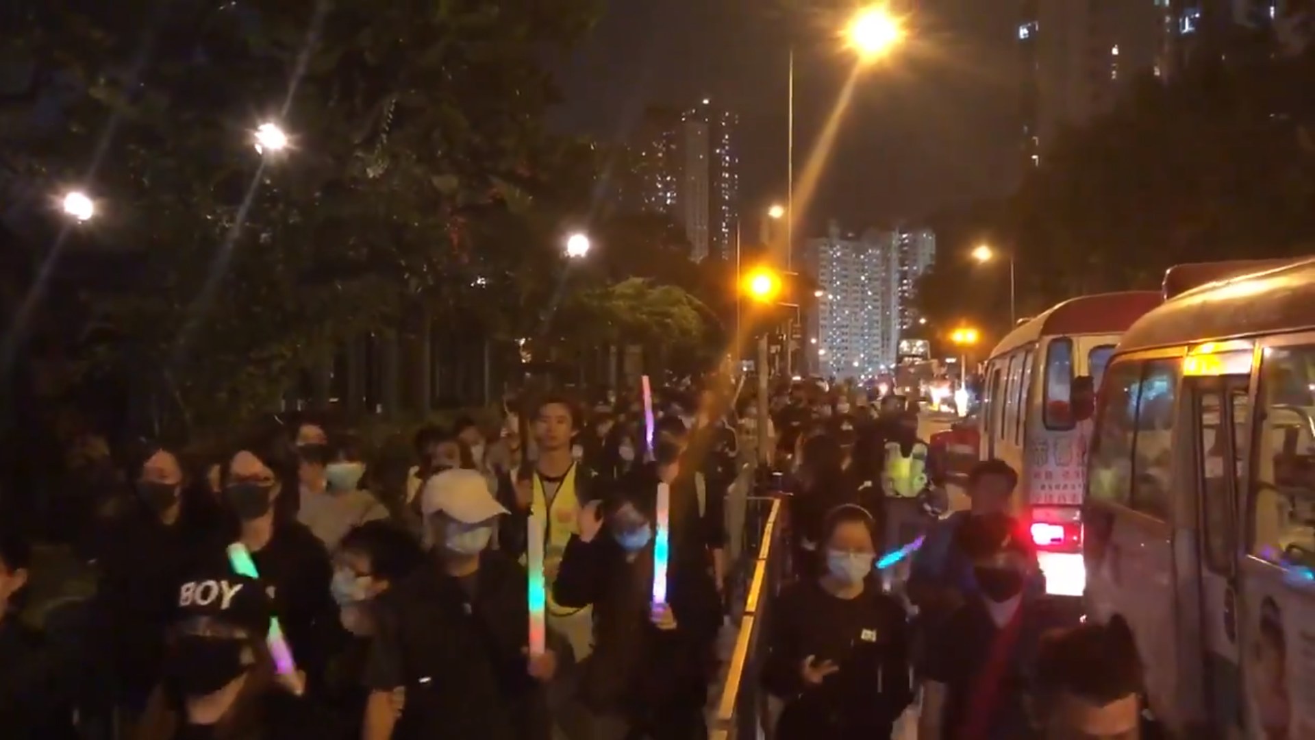 Eine Gruppe von Menschen geht nachts eine beleuchtete Straße entlang, einige tragen Masken, mit Polizeifahrzeugen auf der rechten Seite und Gebäuden und Bäumen, die den Weg säumen.