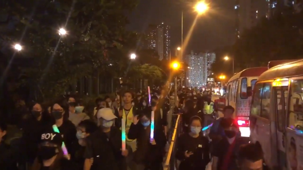 Eine Gruppe von Menschen geht nachts eine beleuchtete Straße entlang, einige tragen Masken, mit Polizeifahrzeugen auf der rechten Seite und Gebäuden und Bäumen, die den Weg säumen.