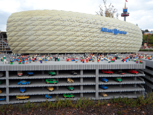 Eine gro├če Lego-Struktur, die der Allianz Arena in M├╝nchen, Deutschland, ├Ąhnelt, mit einer Gruppe von Menschen davor, umgeben von B├Ąumen, Masten, einem Turm und Gr├╝nzeug unten, unter einem bew├Âlktem Himmel.