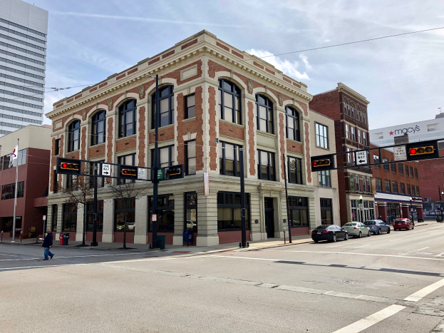 Ein großes Backsteingebäude, das ehemalige Rathaus, steht an der Ecke einer belebten Stadtstraße, umgeben von anderen Gebäuden, Verkehr, Fußgängern und Bäumen unter einem bewölkten Himmel.